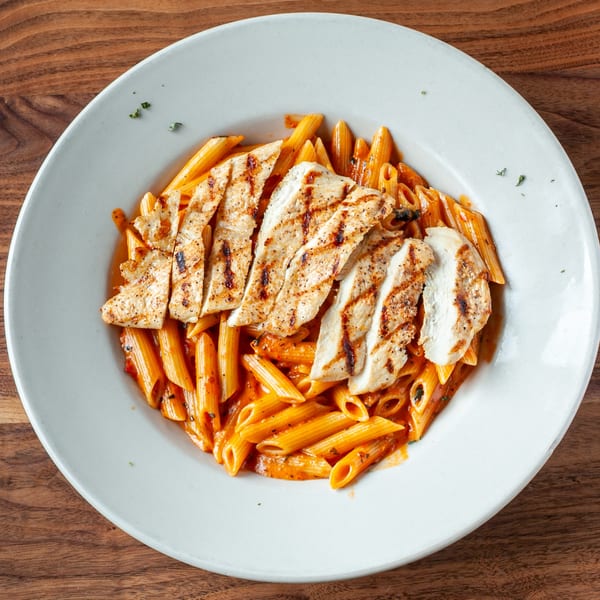 Grilled chicken slices over penne pasta in a creamy red sauce on a white plate.