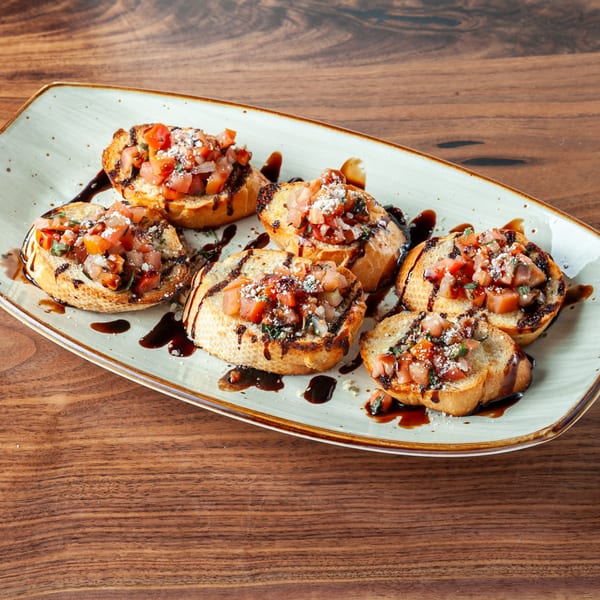 Six pieces of bruschetta with diced tomatoes, cheese, and balsamic glaze on a white plate.