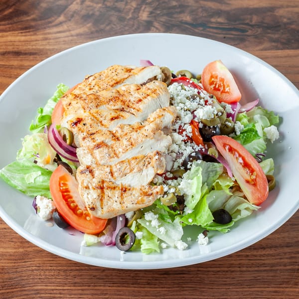 Grilled chicken salad with lettuce, tomatoes, olives, red onion, and feta cheese on a white plate.